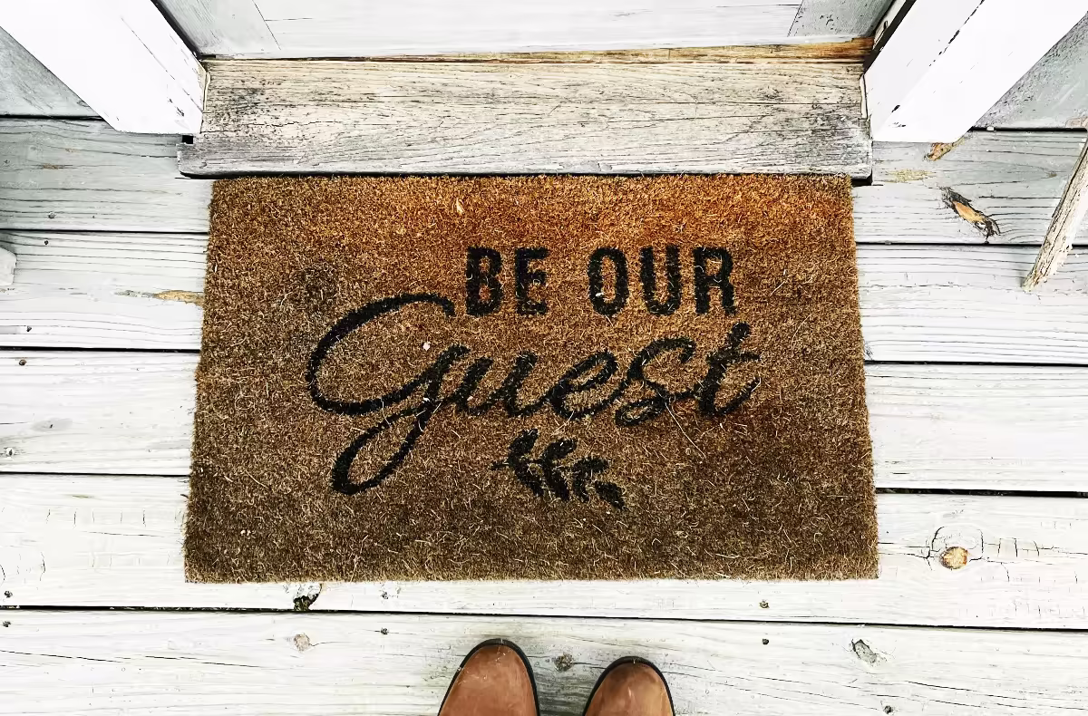 Preș de intrare cu mesajul „Be our guest” pe o verandă din lemn